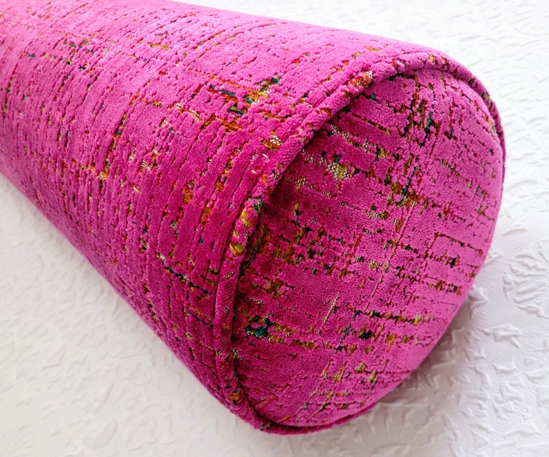 May include: A vibrant magenta bolster pillow with a textured, patterned fabric. The cylindrical pillow features rounded ends and a decorative trim. The fabric has flecks of yellow, green, and blue.