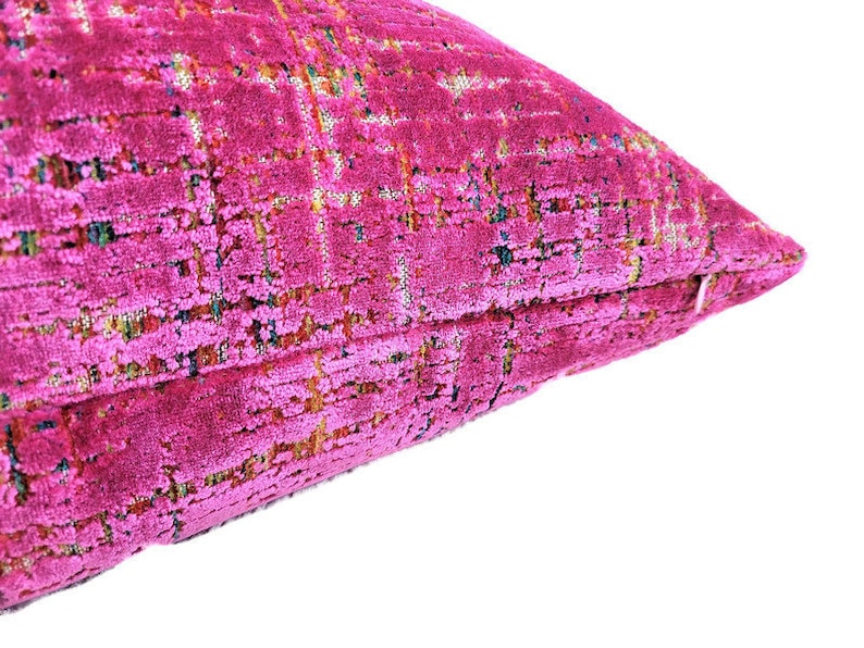 May include: Close-up of a fuchsia-colored decorative pillow. The pillow has a textured, woven appearance with a grid-like pattern. The fabric has a plush, velvety feel, and the color is vibrant and eye-catching.