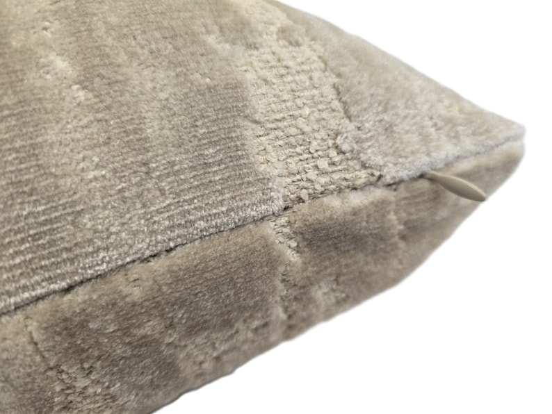 May include: Close-up of a beige velvet pillow with a zipper closure. The pillow has a textured surface and a subtle pattern.