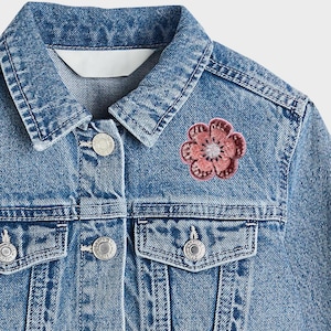 May include: Light blue denim jacket with a flower patch on the chest. The jacket has a classic design with button closures, chest pockets, and a collar. The flower patch is a pink and red color. The jacket is a children's clothing item.