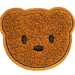 May include: A brown teddy bear patch with a textured, plush appearance. The bear's face features black eyes and a small black nose. The patch is outlined with a dark brown border.
