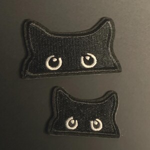 May include: Two black embroidered patches shaped like cat heads with pointed ears. Each patch features two white circular eyes peering out from the bottom edge. The patches are identical in design but differ in size, with one being larger than the other.