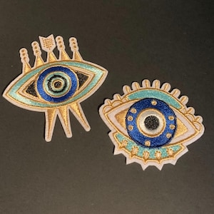 May include: Two embroidered patches featuring stylized eye designs. The left patch has a gold, light blue, and dark blue color scheme with pointed rays. The right patch is circular with a dark blue iris, light blue accents, and gold rays.