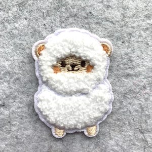 May include: A white, fluffy sheep patch with a detailed embroidered design. The patch features a sheep with a light brown face, ears, and feet, and a smiling expression. The patch is likely for clothing or accessories.