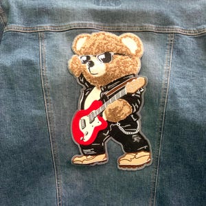 May include: A large embroidered patch featuring a brown teddy bear wearing black sunglasses, a black leather jacket, and black pants, while playing a bright red electric guitar. The patch is attached to the back of a blue denim jacket.