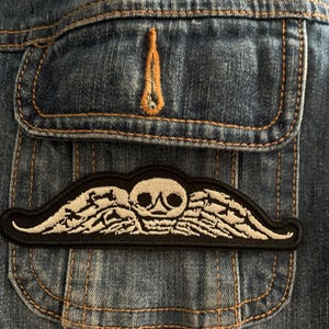 May include: Close-up of a denim jacket pocket with a black and white embroidered patch. The patch features a skull with wing-like designs. The jacket has visible stitching and a small, decorative orange stitch.