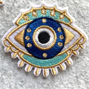 May include: Embroidered patch featuring an eye design. The patch has a white base with gold, blue, and teal accents. The eye's iris is blue with a black pupil, surrounded by gold detailing. The patch is likely for clothing or accessories.