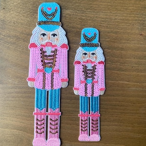 May include: Two decorative nutcracker figures. The figures are in a pink, blue, and brown color scheme. The nutcrackers have a traditional design with a hat, jacket, and boots. The figures are made of a textured material.