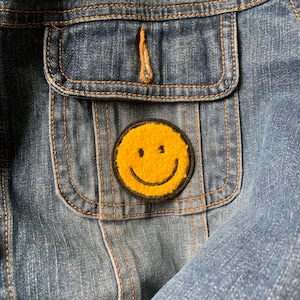 May include: A yellow smiley face patch with a black outline sewn onto a blue denim jacket.