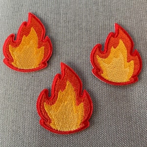 May include: Three embroidered patches shaped like flames, each featuring a bright yellow center surrounded by a vibrant orange and red border. The flame-shaped patches have a textured, stitched appearance with clearly defined edges and a consistent design.