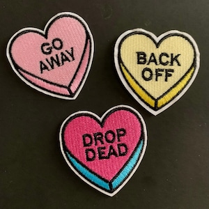 May include: Three heart-shaped embroidered patches. One pink heart says "GO AWAY" in white. A yellow heart says "BACK OFF" in black. A pink heart with a blue border says "DROP DEAD" in white. Each patch has a white border.