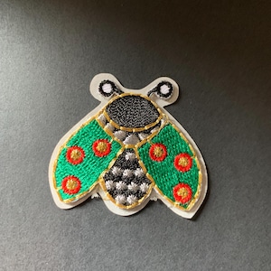 May include: Embroidered patch featuring a stylized insect design. The insect has green wings with red and gold accents, a black and silver body, and gold trim. The patch is on a white backing.