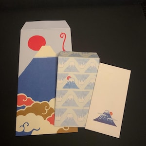 May include: Three paper envelopes featuring designs of Mount Fuji. The largest envelope displays a large blue mountain with a red sun and stylized clouds. The middle envelope has a repeating mountain pattern. The smallest envelope shows a single mountain.
