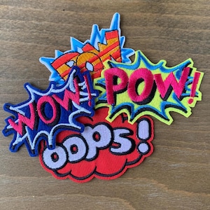 May include: A collection of three embroidered comic book style patches. One patch features the word WOW in pink on a blue starburst. Another displays the word POW in pink on a lime green starburst. The third shows OOPS in black on a red cloud.