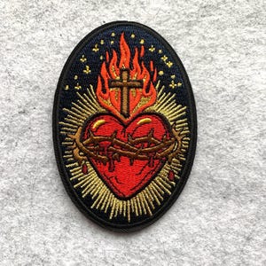 May include: Embroidered patch featuring a red heart with a crown of thorns, a cross, and flames. The patch has a black border and gold accents, set against a navy blue background with small gold stars.