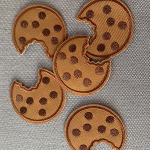 May include: Five circular embroidered patches shaped like cookies with bites taken out of them. Each patch is tan with dark brown embroidered chocolate chips and a stitched border. The patches are arranged in a scattered pattern on a gray surface.
