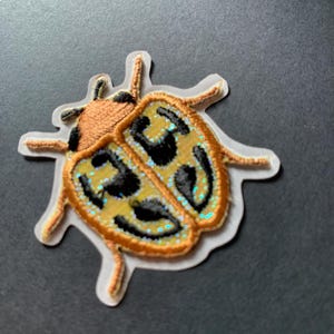 May include: Embroidered ladybug patch with a tan body, black spots, and a yellow and black patterned shell. The patch has a white border and is on a dark gray surface. The ladybug's shell has iridescent glitter accents.
