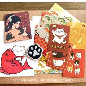 Puede incluir: Una colecci&oacute;n de art&iacute;culos de tem&aacute;tica japonesa, que incluye tarjetas con ilustraciones de gatos de la suerte, un parche con una huella de pata y una ilustraci&oacute;n de pez rojo. La imagen tambi&eacute;n presenta una tarjeta con una mujer y gatos negros.