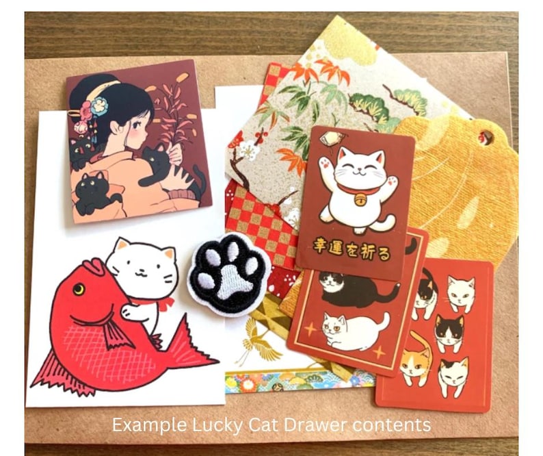 Puede incluir: Una colecci&oacute;n de art&iacute;culos de tem&aacute;tica japonesa, que incluye tarjetas con ilustraciones de gatos de la suerte, un parche con una huella de pata y una ilustraci&oacute;n de pez rojo. La imagen tambi&eacute;n presenta una tarjeta con una mujer y gatos negros.