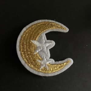 May include: Embroidered patch featuring a crescent moon and two stars. The moon is gold with a white border, and the stars are white. The patch is likely for clothing or accessories.