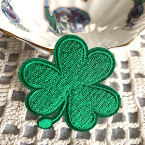 May include: A bright green embroidered shamrock patch with a white border. The shamrock has three distinct leaves and a small stem. The patch is set against a white crocheted background, with a decorative ceramic dish in the background.