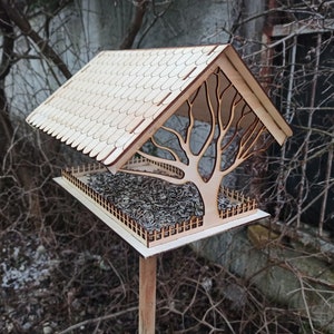 Puede incluir: Un comedero para pájaros de madera con un tejado inclinado y un recorte en forma de árbol. El comedero está lleno de semillas y tiene una pequeña valla alrededor del área de alimentación. Está montado en un poste de madera para atraer aves.