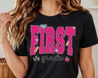May include: A black short-sleeve t-shirt featuring the text FIRST in large pink block letters with white polka dots. Below, the word grade is written in a cursive black font. Small school-themed icons like a pencil, apple, and paper plane are included.