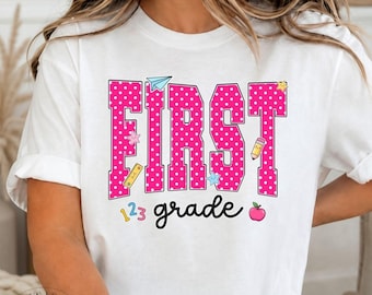 May include: A white short-sleeve t-shirt featuring the text FIRST in large, pink, polka-dotted block letters and the word grade in black cursive script. The design includes small colorful school-themed icons like a pencil, apple, and numbers.