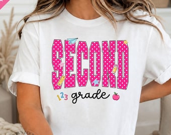 May include: A white short-sleeved t-shirt featuring the word SECOND in large, pink, white-polka-dotted letters, with the word grade written below in black script. The design includes small colorful icons of a paper airplane, pencil, and apple.