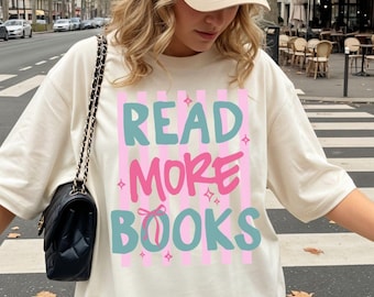 May include: A beige baseball cap with a small dark logo and an oversized cream-colored t-shirt featuring the text READ MORE BOOKS in teal and pink letters over vertical pink stripes, paired with a black quilted shoulder bag with a gold chain strap.