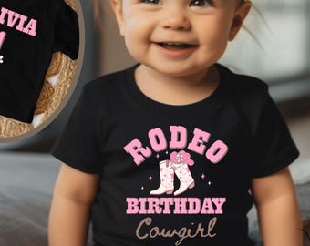 May include: A black t-shirt with the text "RODEO BIRTHDAY Cowgirl" in pink, featuring a graphic of cowboy boots. A second black t-shirt with the name "OLIVIA" and the number "1" in pink is also shown. The image is of a baby wearing the t-shirt.