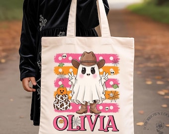 May include: A cream-colored canvas tote bag featuring a graphic of a ghost wearing a brown cowboy hat and boots, making a peace sign. The design includes orange and pink stripes, pumpkins, and spiderwebs, with the name OLIVIA printed in pink.