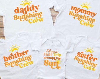 May include: A set of five white t-shirts and one white baby bodysuit featuring orange and yellow sun graphics and text. The shirts read daddy Sunshine Crew, mommy Sunshine Crew, brother Sunshine Crew, sister Sunshine Crew, and Olivia's First Trip around the Sun.