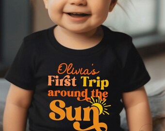 May include: A black short-sleeved t-shirt featuring the text "Olivia's First Trip around the Sun" in orange and yellow lettering, accompanied by a graphic of a sun. The design uses a retro-inspired font style across the chest of the garment.