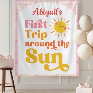 May include: A banner with the text "Abigail's First Trip around the Sun" in pink, orange, and yellow. A smiling sun graphic is included. The banner is framed with a pink plaid pattern. Balloons and a gift box are also in the image.