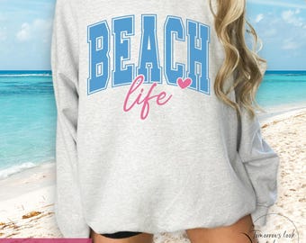 May include: A light gray sweatshirt with the words "BEACH life" in blue and pink lettering. The word "BEACH" is in a block letter style. The word "life" is in a cursive font. A pink heart is between the words.