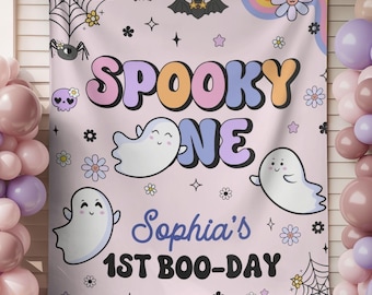 May include: A light pink fabric backdrop featuring the text SPOOKY ONE and Sophia's 1ST BOO-DAY. The design includes cartoon ghosts, spider webs, bats, flowers, and a rainbow. The banner is decorated with clusters of pink and purple balloons.