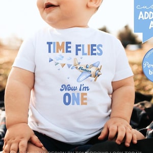 May include: A white short-sleeved shirt with a blue airplane graphic and the text "TIME FLIES Now I'm ONE". The airplane has a blue and brown design with the name "Gludson" written on it.