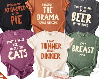 May include: Six short-sleeved t-shirts in black, orange, charcoal, purple, cream, and green, each featuring unique text designs. A hand holds a plaid can cooler with the text FRIENDSGIVING 2026 THE CREW CHEERS. The shirts display various humorous phrases.