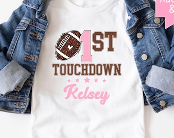 May include: A white t-shirt featuring a brown glitter football graphic, the text 1ST TOUCHDOWN, and the name Kelsey in pink script. The shirt is layered under a blue denim jacket. A pink speech bubble graphic includes the text custom Add Name & Age.