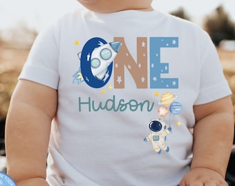 May include: A white short-sleeved t-shirt featuring the word ONE in large blue and brown letters, with a rocket ship graphic inside the letter O. The name Hudson is printed below in teal, alongside an astronaut holding planet-shaped balloons.
