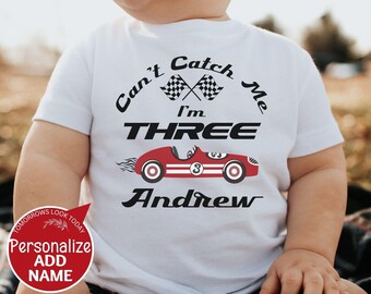 May include: A white short-sleeved t-shirt featuring a graphic of a red race car with the number 3, two checkered flags, and the black text "Can't Catch Me I'm THREE Andrew". A red circular badge with white text "Personalize ADD NAME" is visible.