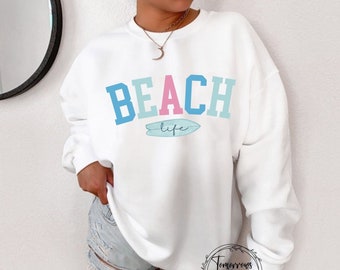 May include: White sweatshirt with the text "BEACH life" in blue, pink, and light blue letters with a surfboard graphic underneath.