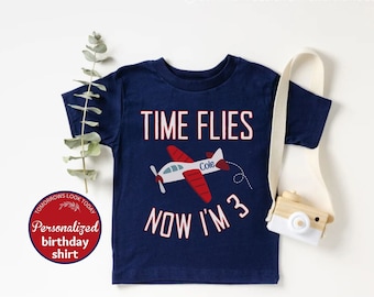 May include: Navy blue short-sleeved t-shirt with the text "TIME FLIES NOW I'M 3" and a red and white airplane graphic. The shirt includes the name "Cole" on the airplane. A personalized birthday shirt.