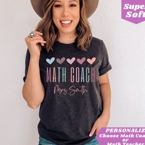 May include: A dark grey t-shirt with the text "MATH COACH" in rainbow colors and "Mrs. Smith" in pink. The shirt has five hearts above the text, one with a leopard print and the rest in various shades of pink and blue.