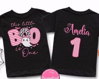 May include: Two black t-shirts. The left shirt features a pink striped graphic with the text "this little BOO is One" and a ghost with a leopard print bow. The right shirt displays the name "Amelia" in pink script above the number "1".