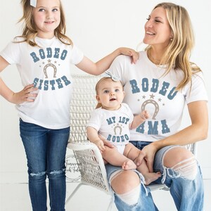 May include: A family of three wearing white t-shirts with blue text and a horseshoe graphic. The shirts say "Rodeo Sister", "My 1st Rodeo", and "Rodeo Mama".
