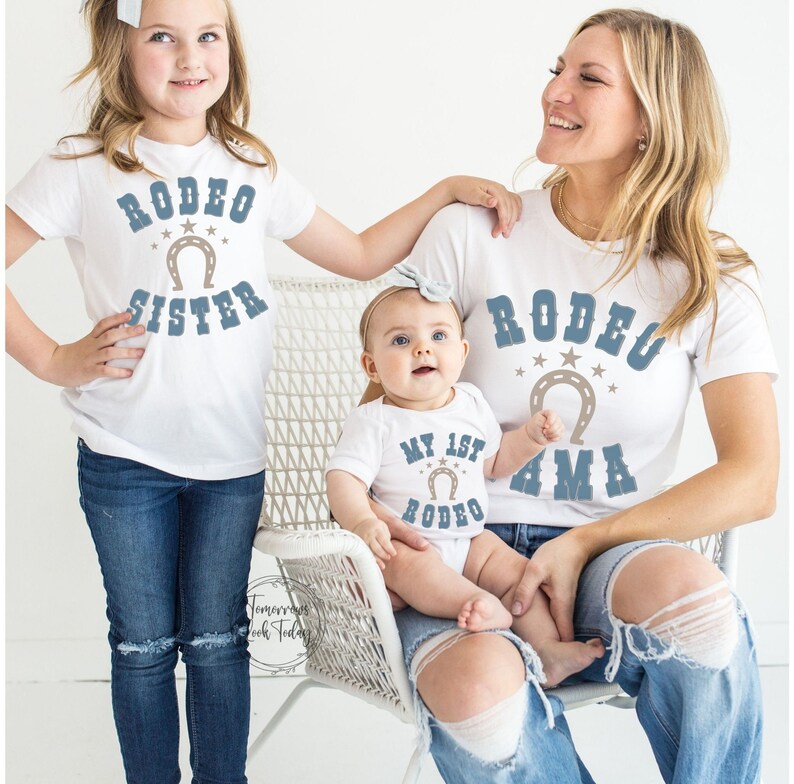May include: A family of three wearing white t-shirts with blue text and a horseshoe graphic. The shirts say "Rodeo Sister", "My 1st Rodeo", and "Rodeo Mama".