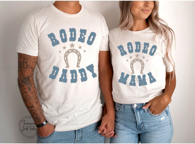 May include: Two white t-shirts with blue text and a horseshoe graphic. The text on the first shirt says "Rodeo Daddy" and the text on the second shirt says "Rodeo Mama".