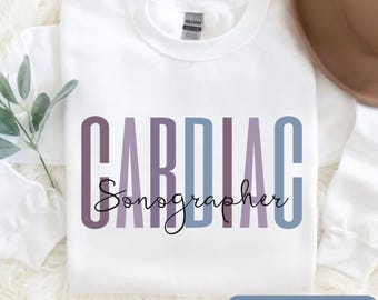 May include: White crew neck sweatshirt with the words "CARDIAC Sonographer" in purple and blue lettering. The sweatshirt also has a small tag that says "GILDAN". The bottom right corner of the image has a button that says "MORE COLORS AVAILABLE".